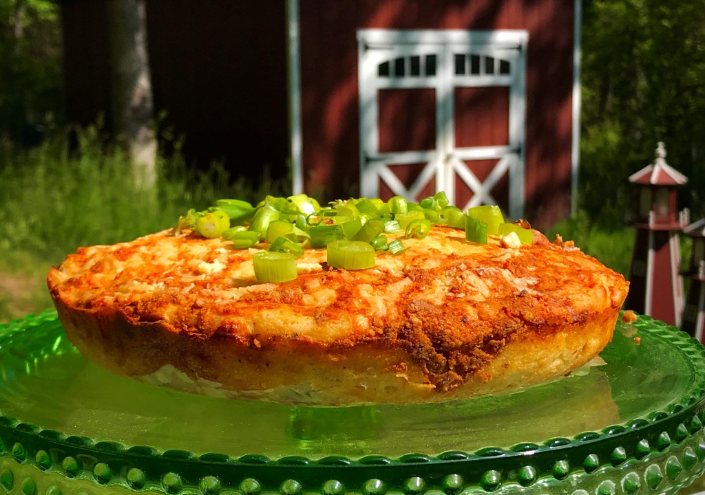 A delicious looking quiche that is set in front of a red barn. The quiche has cut scallions on top. 